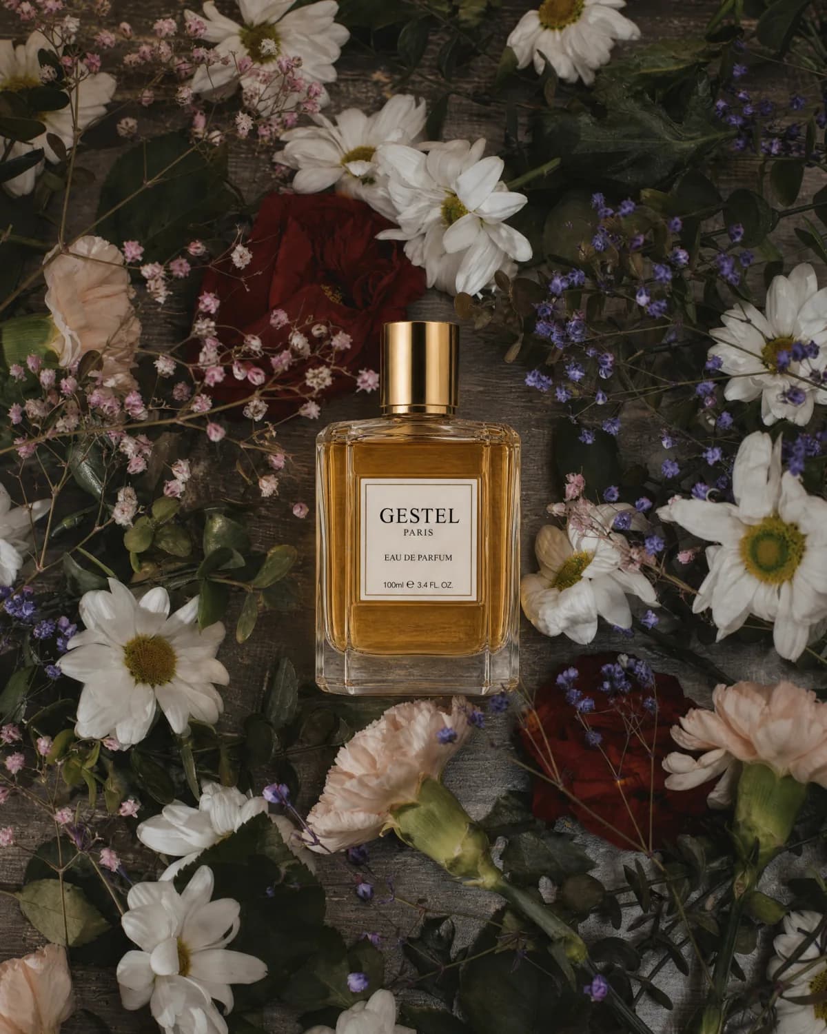 GESTEL perfume bottle surrounded by daisies, roses, and small flowers on a dark wood table.